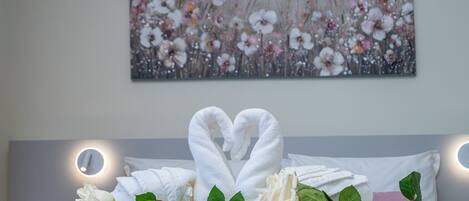 Una cama bien hecha con toallas dispuestas en forma de corazón, una rosa y hojas verdes sobre la cama, con un cuadro floral en la pared de fondo.