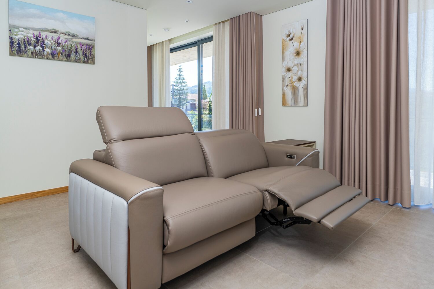 Un sofá reclinable color beige con reposapiés, una mesita auxiliar y un cuadro de flores en la pared.