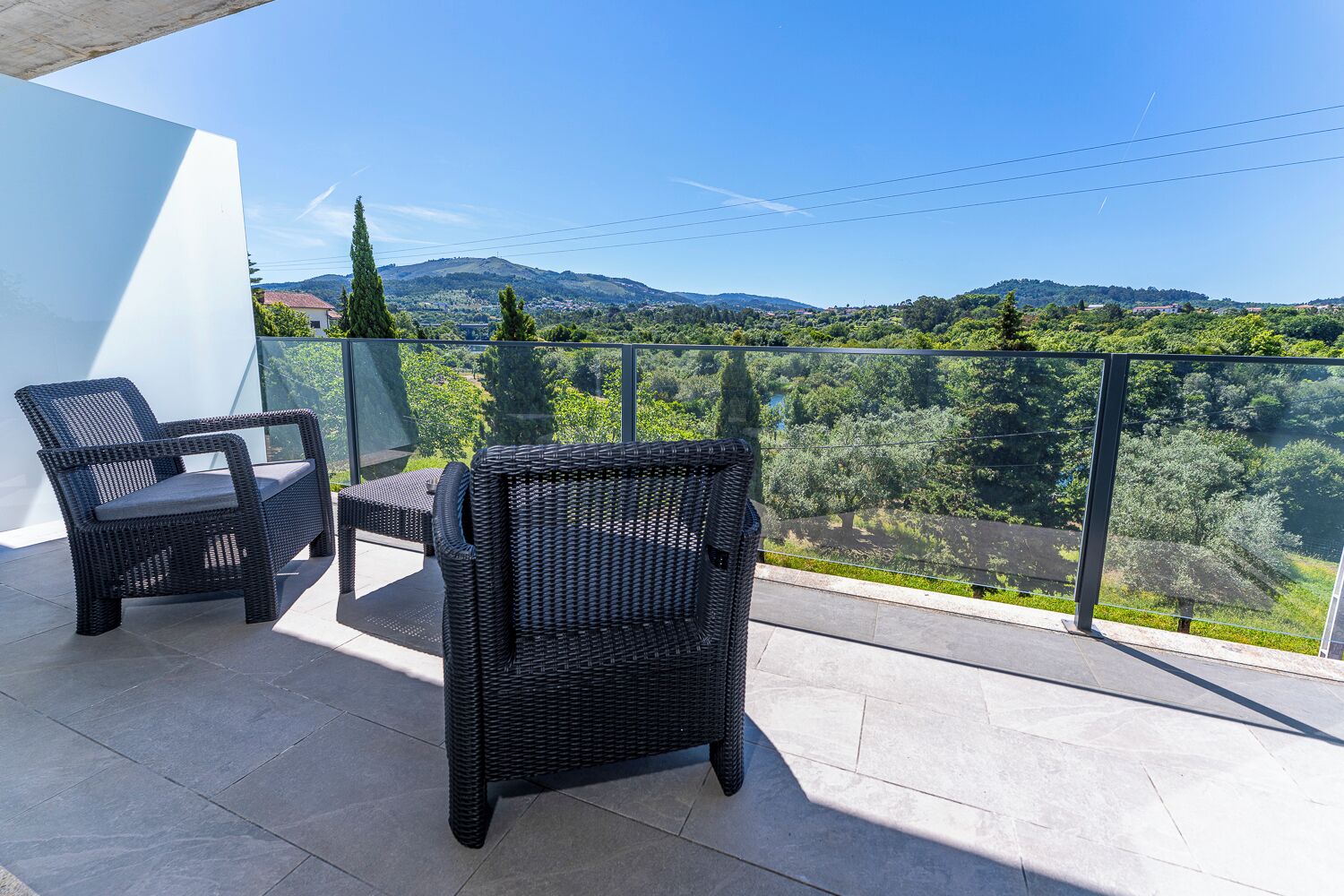 A balcony with wicker furniture and a glass railing overlooking a mountainous landscape.