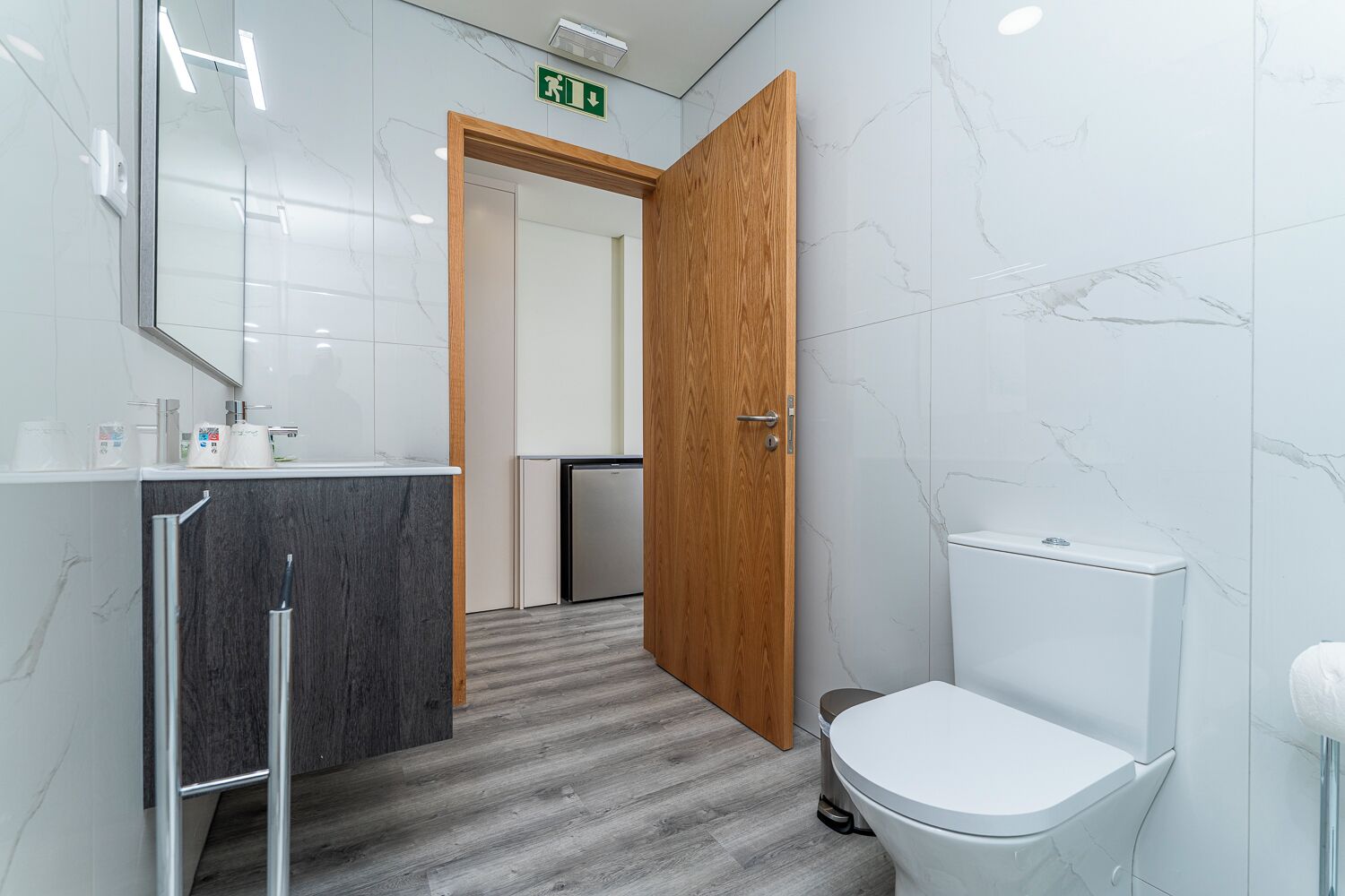 A modern bathroom with a marble wall, a wooden door, a white toilet, and a sink with a mirror.