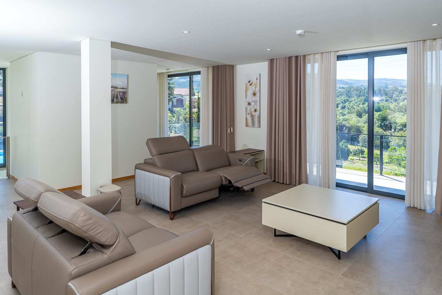 Una sala de estar moderna con un sofá, un sillón reclinable, una mesa de centro y vistas a un paisaje verde a través de amplios ventanales.