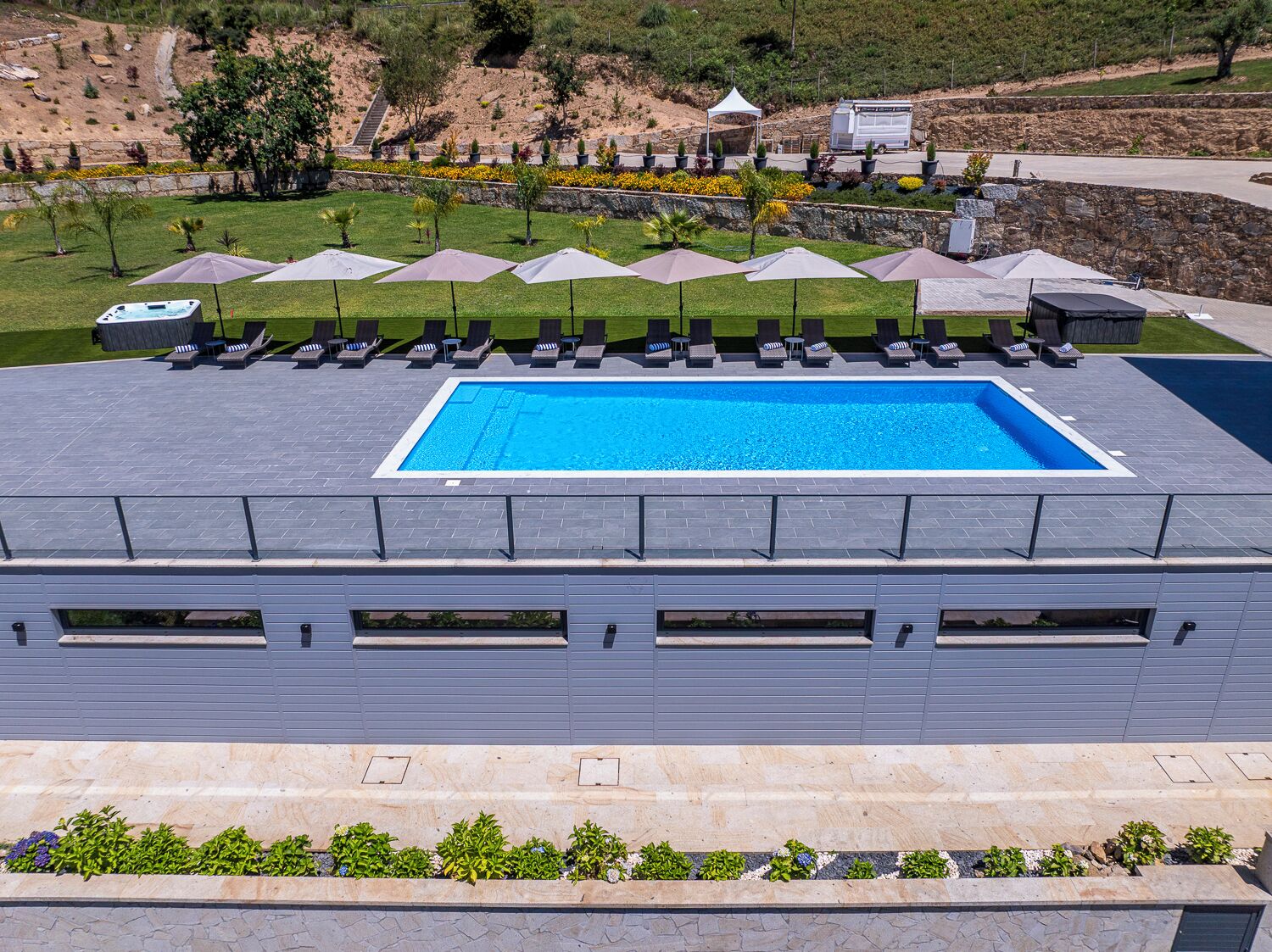 A rooftop pool with lounge chairs and umbrellas, surrounded by a stone wall and greenery.