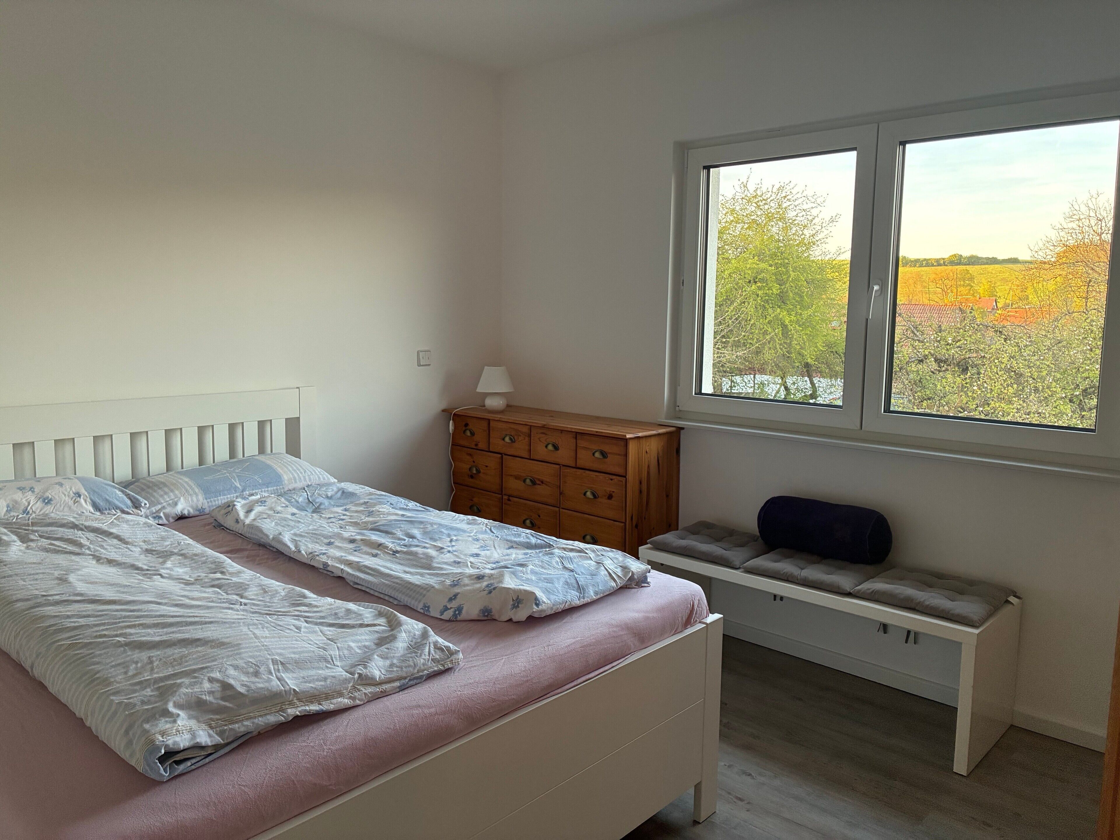 Un dormitorio con cama, cómoda, banqueta y ventana con vista a árboles.