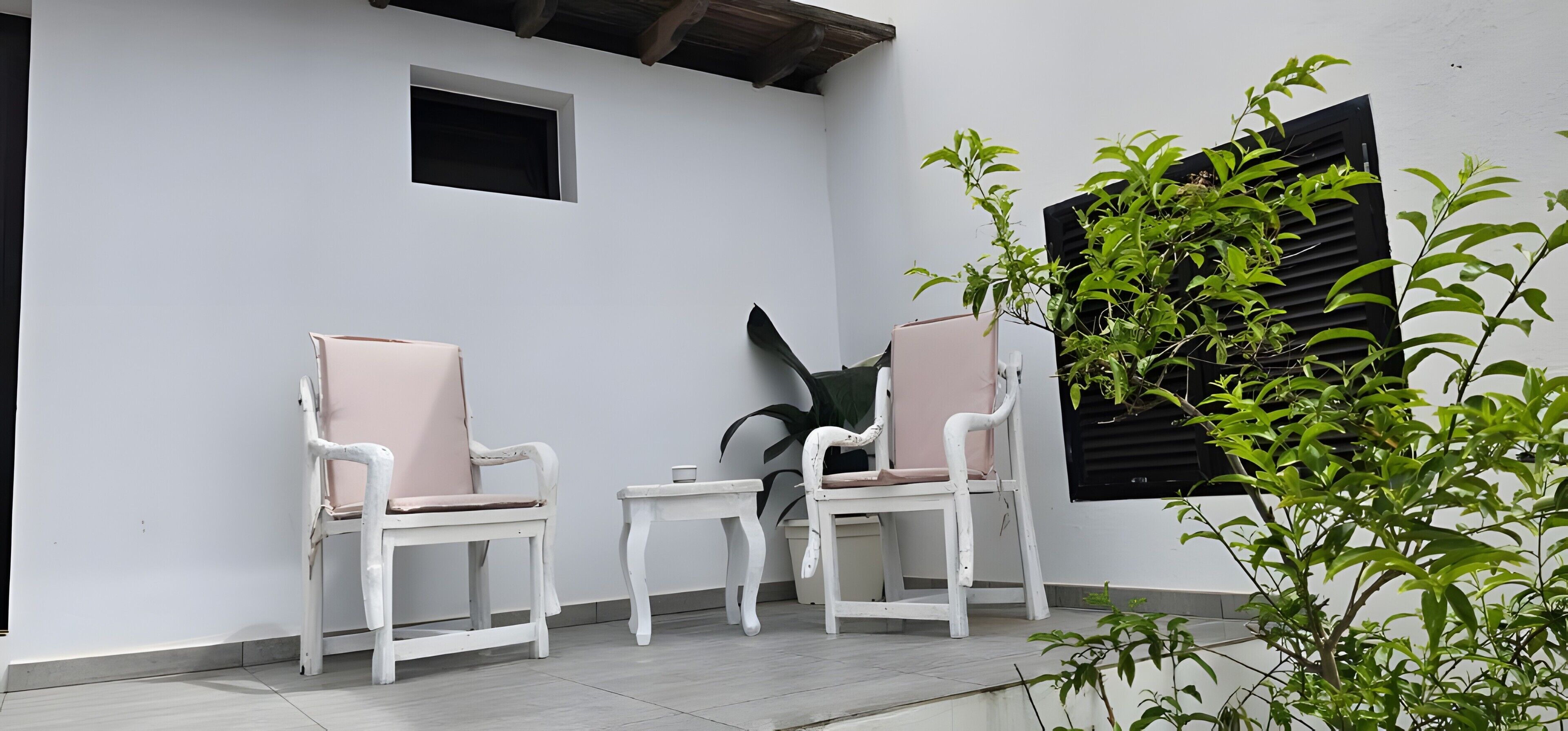 Two white chairs with pink cushions, a small table, and a potted plant in a white-walled outdoor area.