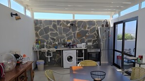 A modern kitchen with stone backsplash, a washing machine, and a dining area with wicker chairs. - Alunita Room – Cozy Retreat in Peaceful Nazaret, Lanzarote (Nazaret)