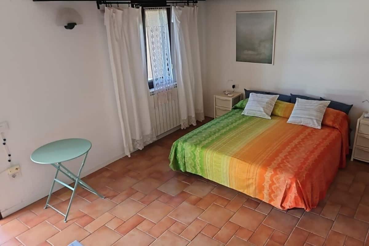 Ein Schlafzimmer mit einem Bett, einem Nachttisch, einem Sessel, einem Fenster mit Vorhängen und einem Wandbild.