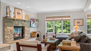 A living room with a stone fireplace, comfortable seating, a television, and large windows with a view of greenery. - Topnotch Luxury Retreat | Sleeps 12 • Pet Friendly • Game Room (Stowe)