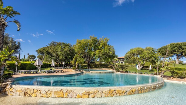 Ein kreisförmiger Pool mit Steinrand, umgeben von einer Terrasse mit Bänken und Sonnenschirmen, mit üppigem Grün im Hintergrund.