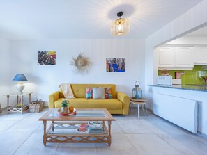 Ein modernes Wohnzimmer mit einem gelben Sofa, einem Glastisch und einer Küchenzeile mit weißen Schränken.