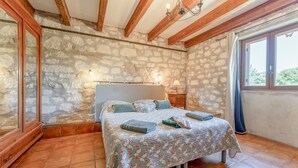 Un dormitorio con pared de piedra, vigas de madera, una cama con colcha estampada, una mesita de noche y una ventana con cortinas.