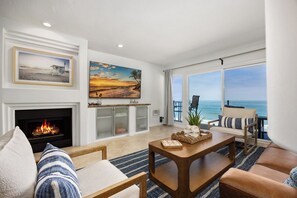 A living room with a fireplace, a large painting, a view of the ocean, and a cozy seating area. - Solana Sunshine! Oceanfront Condo in Solana Beach with Tesla Charger (Solana Beach)