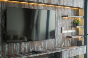A modern living room with a large flat-screen TV mounted on a wooden wall, a small shelf with a plant, and a speaker. - Brand new 3BR Pool Villa with Outdoor Dining (Si Sunthon)