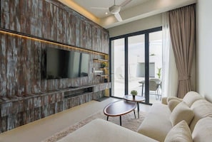 A modern living room with a large flat-screen TV, a sofa, a coffee table, and a sliding glass door leading to an outdoor area. - Brand new 3BR Pool Villa with Outdoor Dining (Si Sunthon)