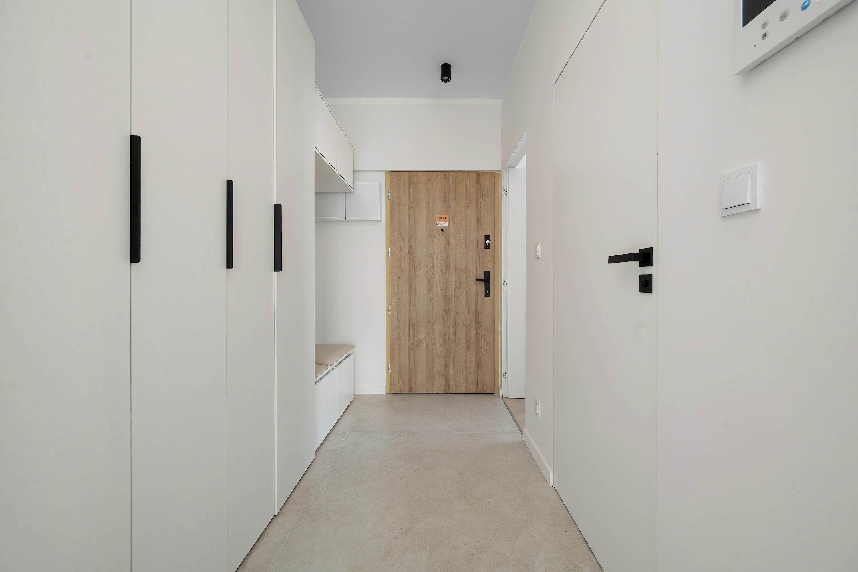 A hallway with white walls, a wooden door at the end, and a light switch on the wall.