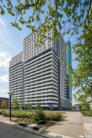 Ein mehrstöckiges Gebäude mit moderner Architektur, das eine Mischung aus Balkonen und Fenstern aufweist und von Grünflächen sowie einem klaren Himmel umgeben ist.