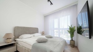 Un dormitorio moderno con cama, mesita de noche, lámpara, planta en maceta y un ventanal grande.