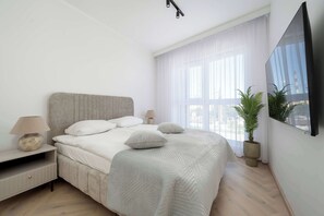 Ein modernes Schlafzimmer mit Bett, Nachttisch, Lampe, Topfpflanze und einem großen Fenster.