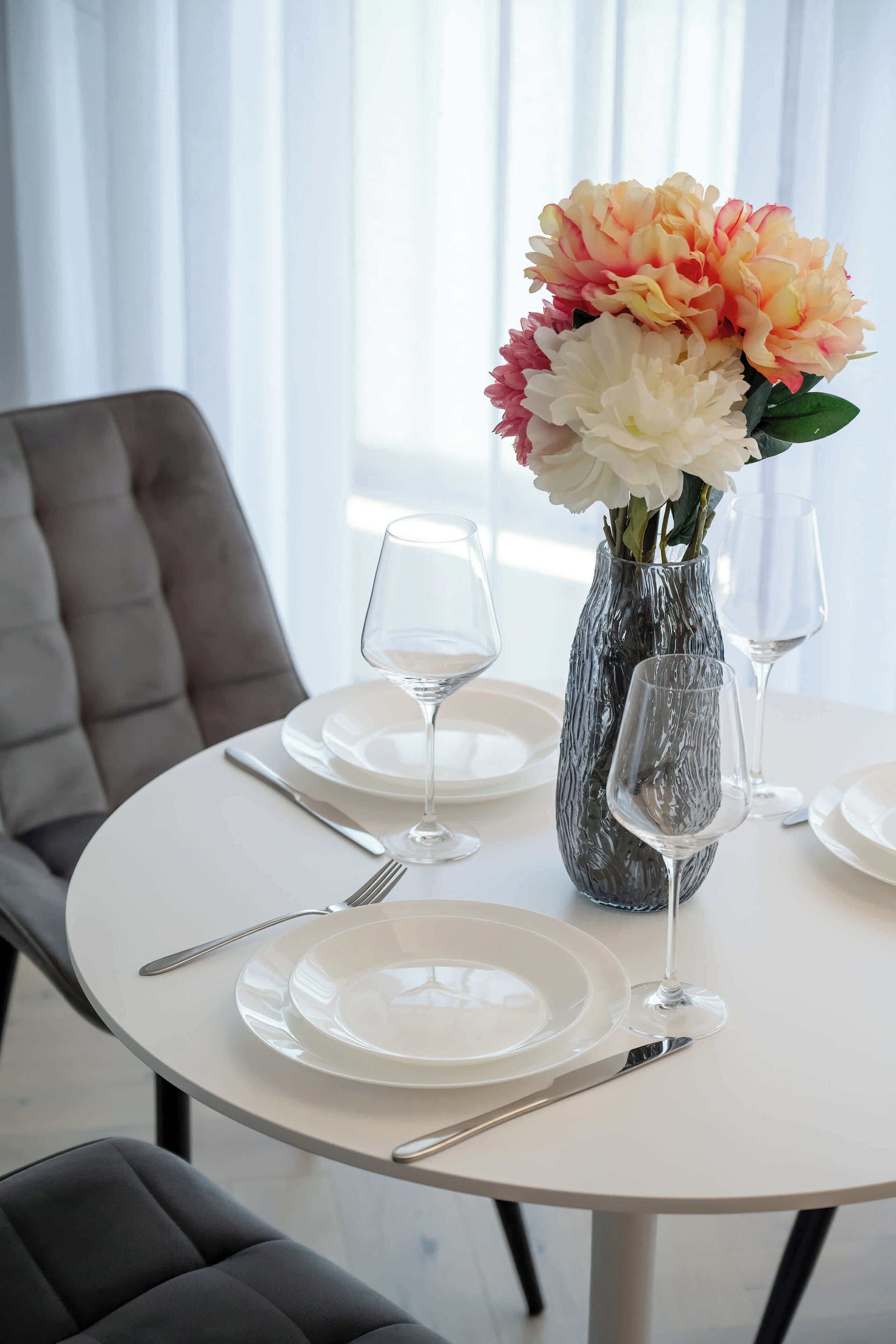 A round dining table with white plates, cutlery, and a vase with flowers.