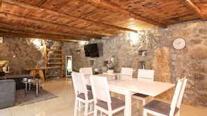A stone-walled room with a wooden ceiling, a dining table with chairs, a television, and a fireplace.