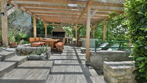 An outdoor patio with stone steps, a wooden pergola, a barbecue grill, and a seating area with cushions.