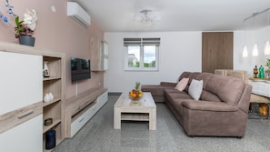 Ein modernes Wohnzimmer mit einem Flachbildfernseher, einem braunen Sofa, einem Couchtisch mit Obst und einem Fenster mit Jalousien.