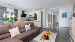 Ein modernes Wohnzimmer mit einem braunen Sofa, einem Couchtisch mit Obst und einer Küche im Hintergrund.