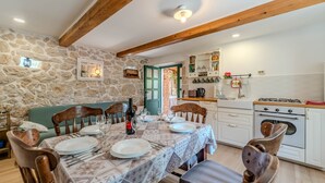 A dining area with a stone wall, wooden beams, a table set for four, and a kitchen with white cabinets and a gas stove. - Guest House Sunset Kalla by Villas Guide (Rab)