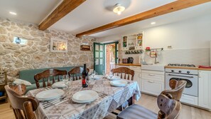 Un coin repas avec un mur en pierre, des poutres apparentes, une table dressée pour quatre personnes et une cuisine dotée de meubles blancs et d’une cuisinière à gaz.