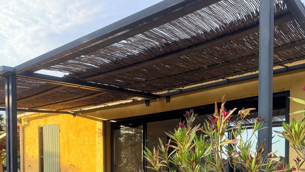 Een moderne buitenruimte met een pergola, een gele muur en een plant met rode bloemen.