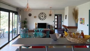 Een moderne woonkamer met een glazen eettafel, smaragdgroene stoelen, een grote wandklok en een televisie.