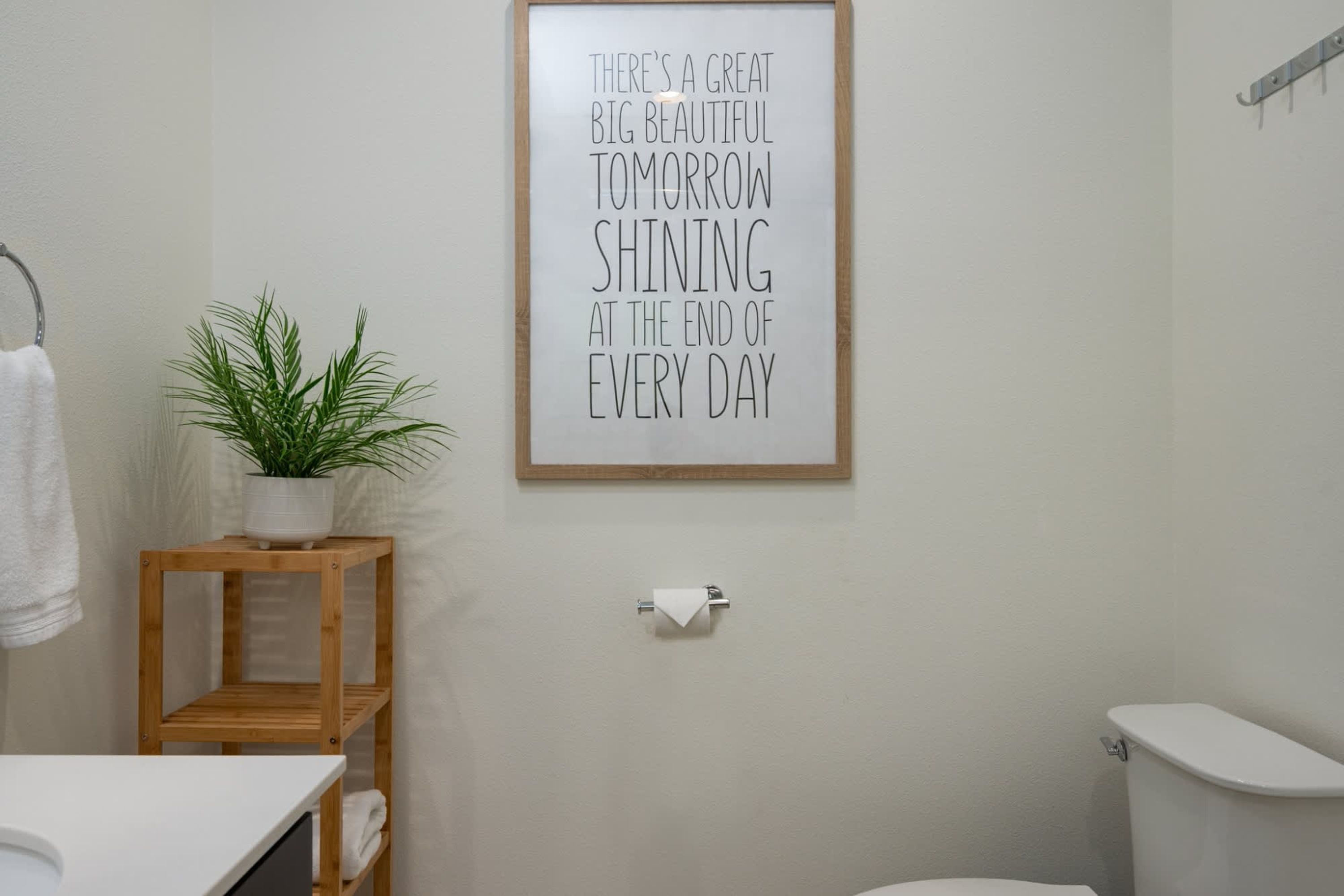 A bathroom with a framed inspirational quote, a wooden shelf with a plant, a toilet, and a towel rack.