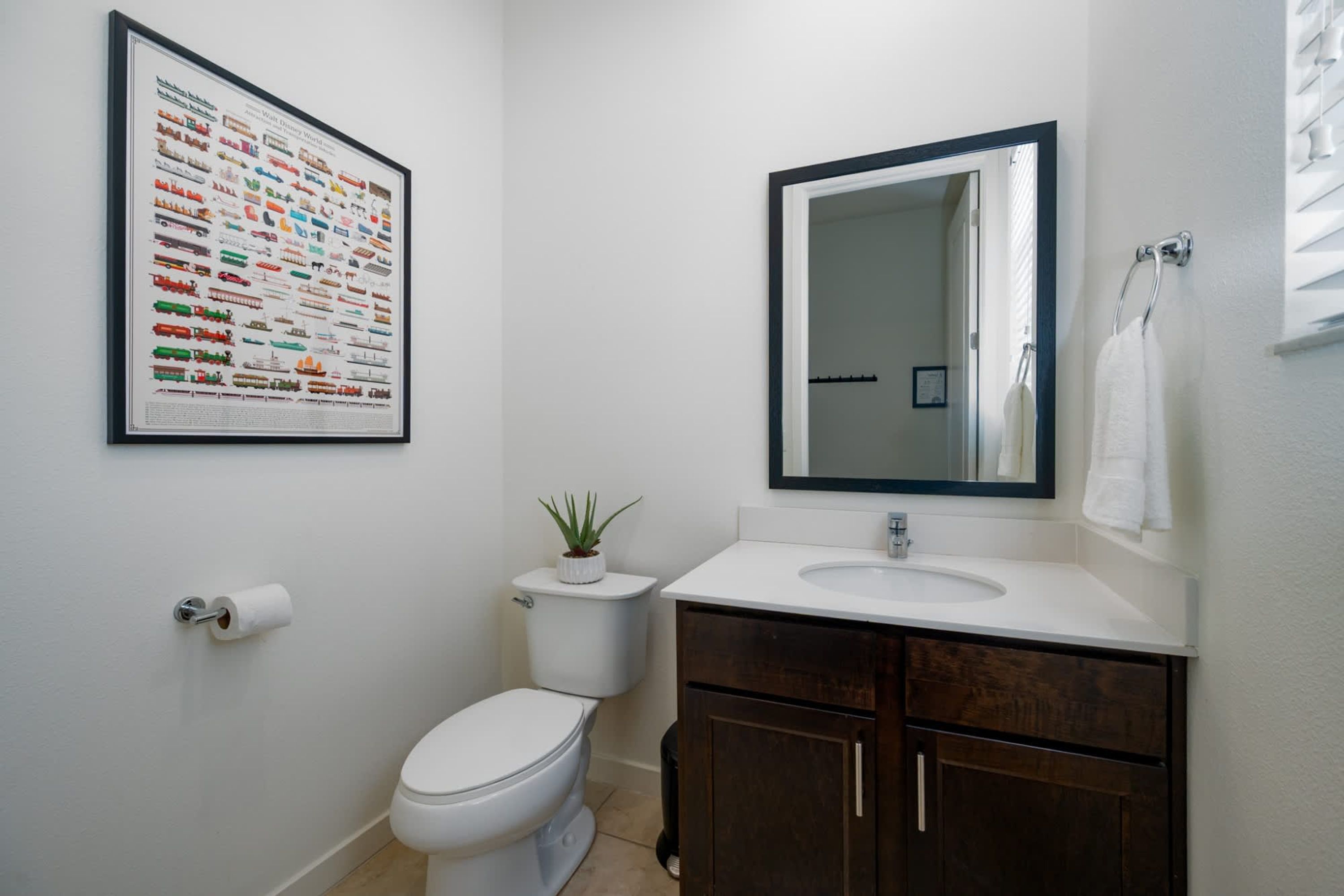 A bathroom with a toilet, sink, mirror, framed picture, and a small plant.