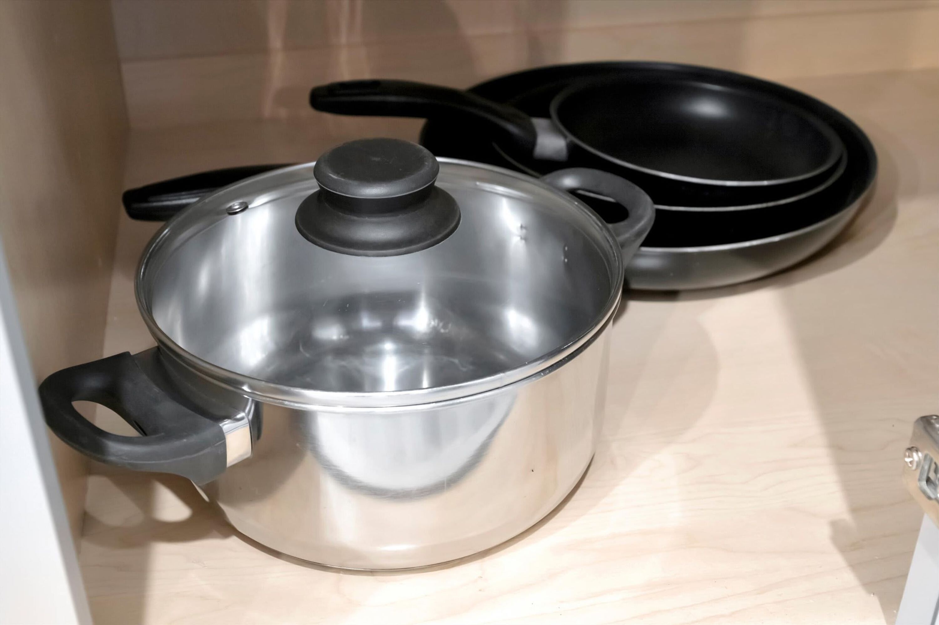 A stainless steel pot with a lid and handles, a black frying pan, and a black skillet on a wooden surface.