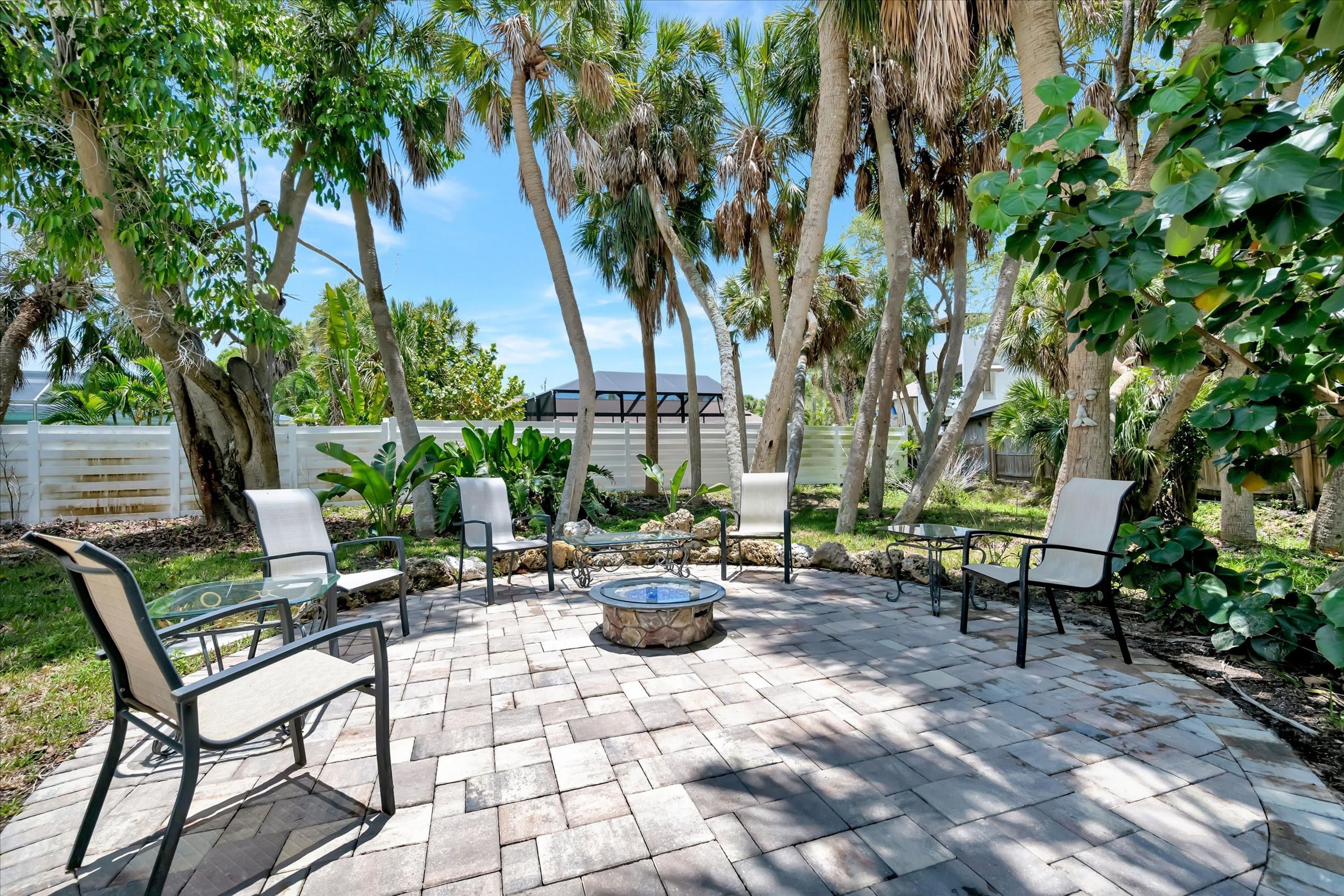 Une terrasse extérieure dotée d’un foyer en pierre, entourée de chaises et de tables, avec en arrière-plan des palmiers et une clôture blanche.