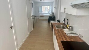 En smal hall leder in till ett sovrum med en säng, ett fönster och en liten köksdel med ett handfat och en spis.