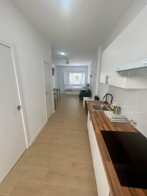 Een smalle gang die leidt naar een slaapkamer met een bed, een raam en een kitchenette met een spoelbak en fornuis.