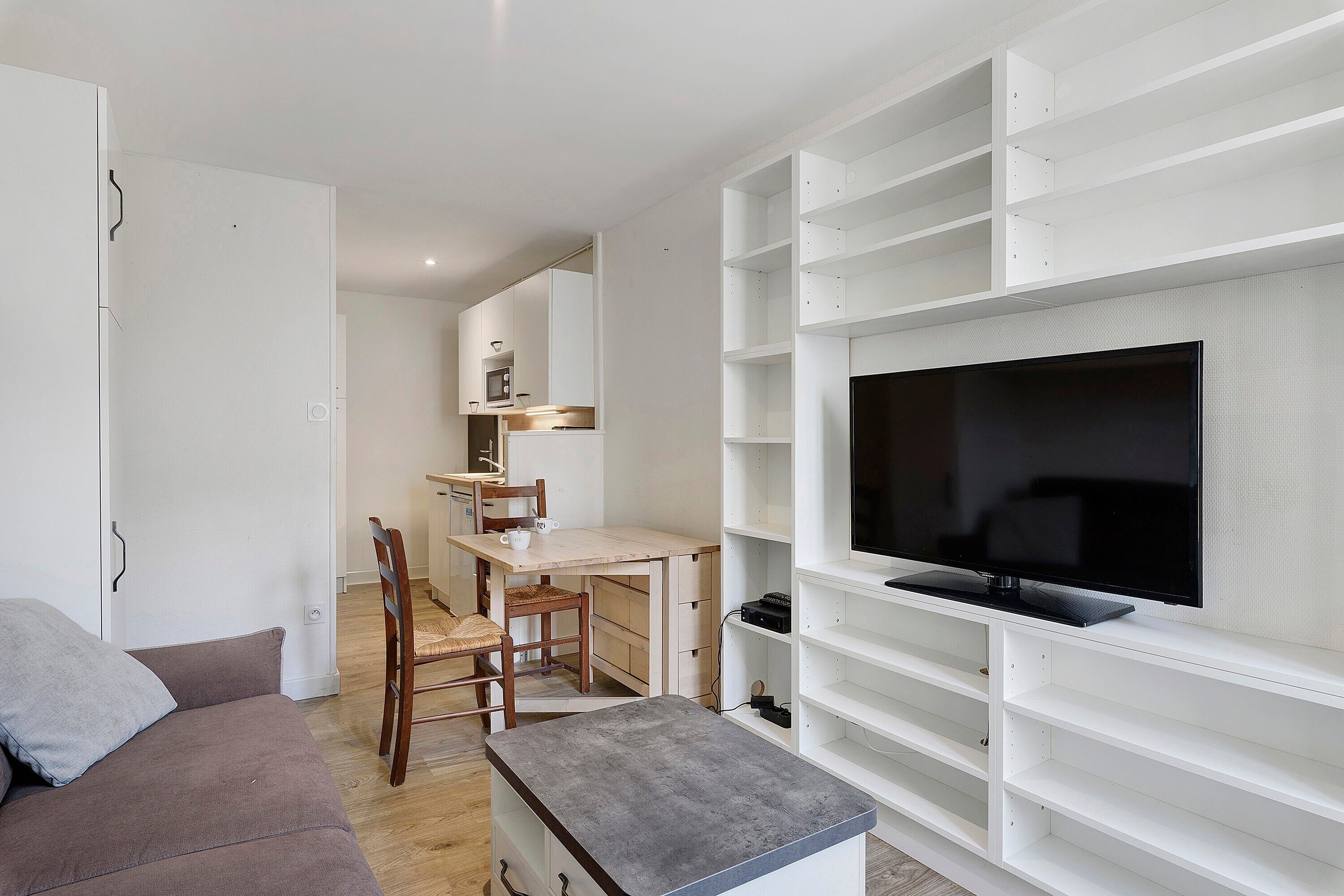 Un salon moderne avec un téléviseur à écran plat fixé sur une étagère blanche, une table à manger en bois avec des chaises et un canapé gris.