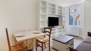 Una sala de estar con una mesa de comedor, sillas, un televisor de pantalla plana, estanterías empotradas y una ventana con vistas a edificios.