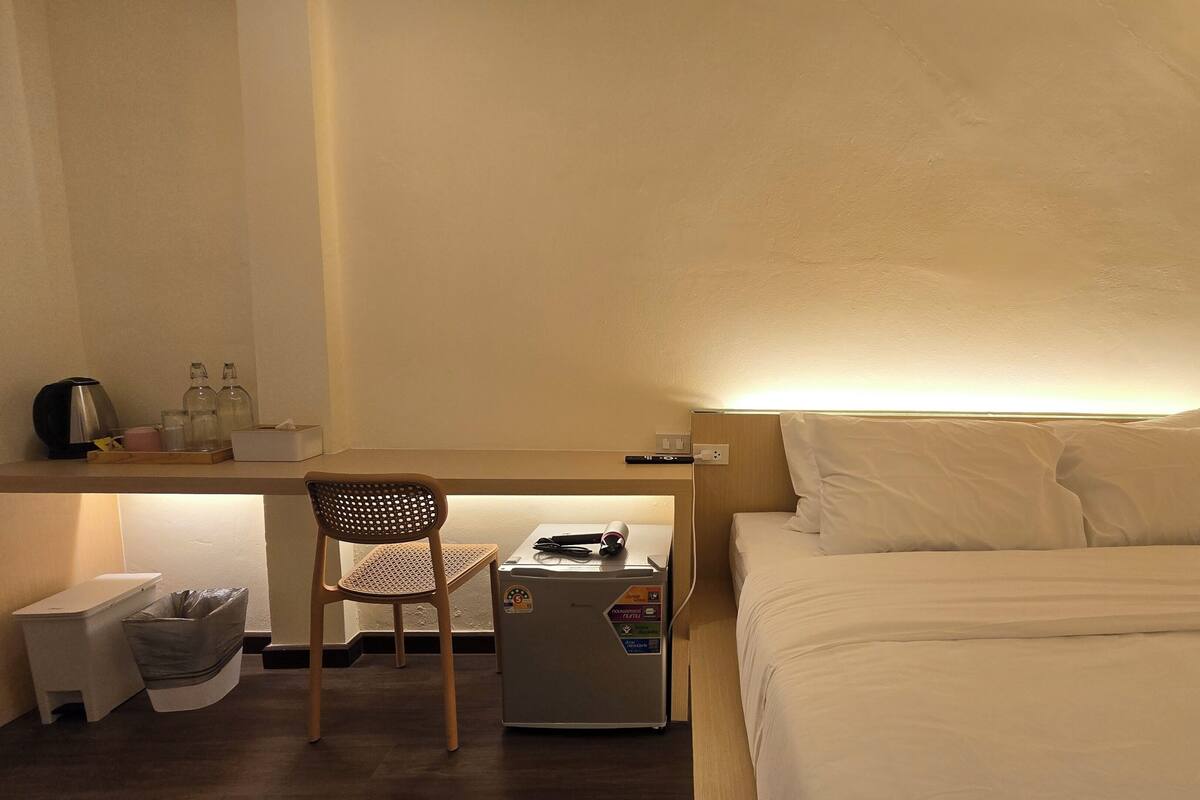 A hotel room with a bed, a desk with a chair, a refrigerator, a trash bin, and a small table with a kettle and glasses.