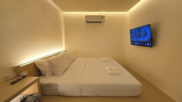 A hotel room with a bed, a flat-screen TV mounted on the wall, and an air conditioning unit above the bed.