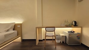 A compact room with a bed, a desk with a chair, a refrigerator, and a small trash bin. - Norns Silom Cafe & Stay (Bangkok)