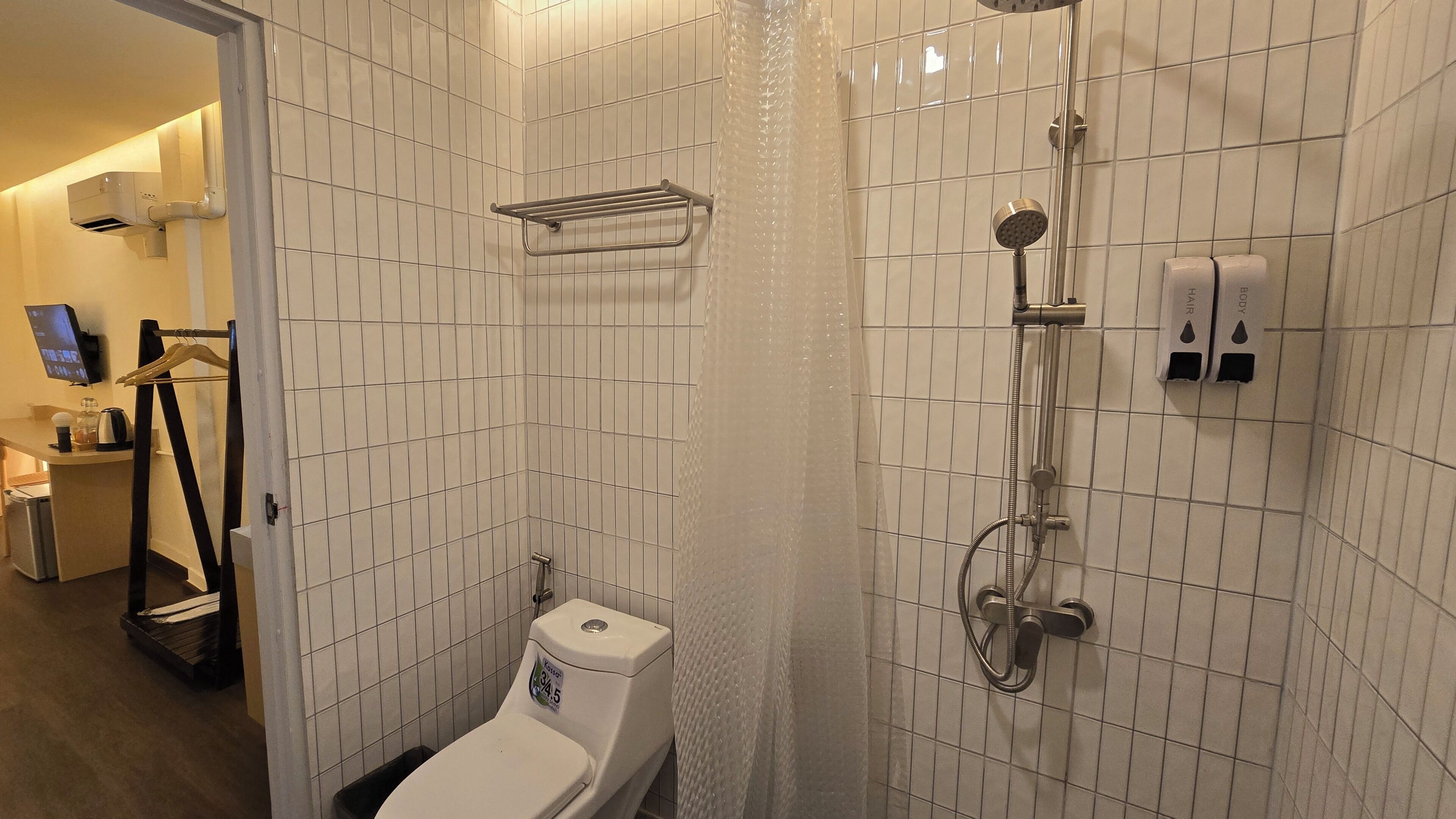 Et badeværelse med hvide flisevægge, et toilet, en bruser med håndbruser og en bøjle til håndklæder.