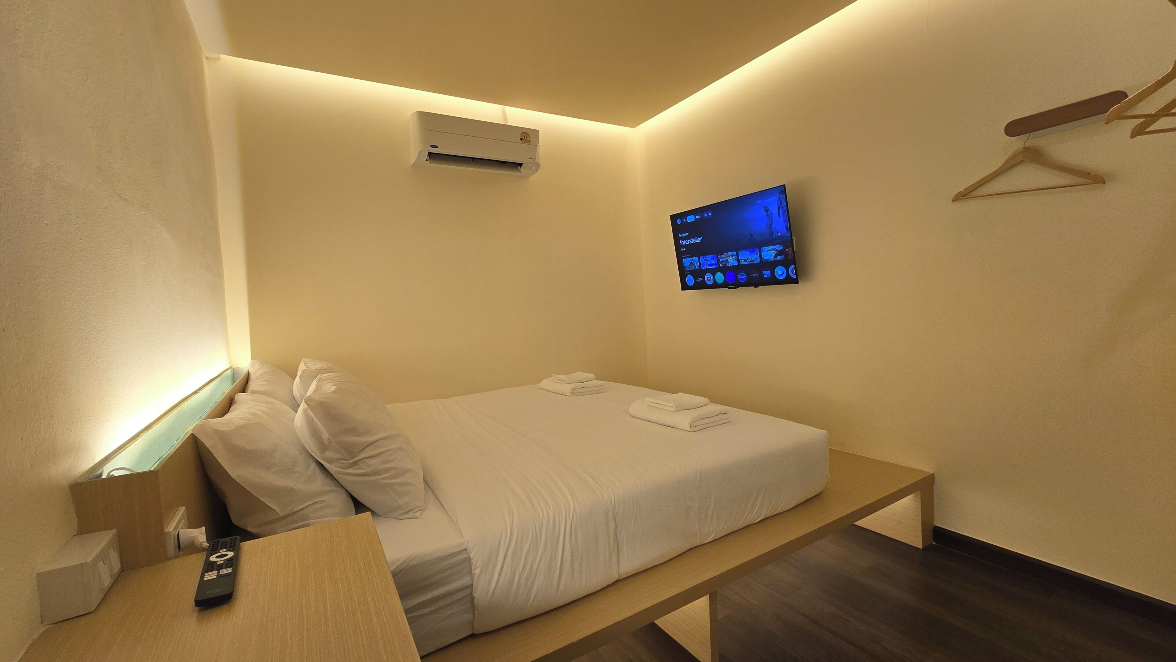 Camera d'albergo di dimensioni ridotte con un letto singolo, un comodino con telecomando, un condizionatore d'aria, una televisione a muro che mostra un programma e una panca in legno.