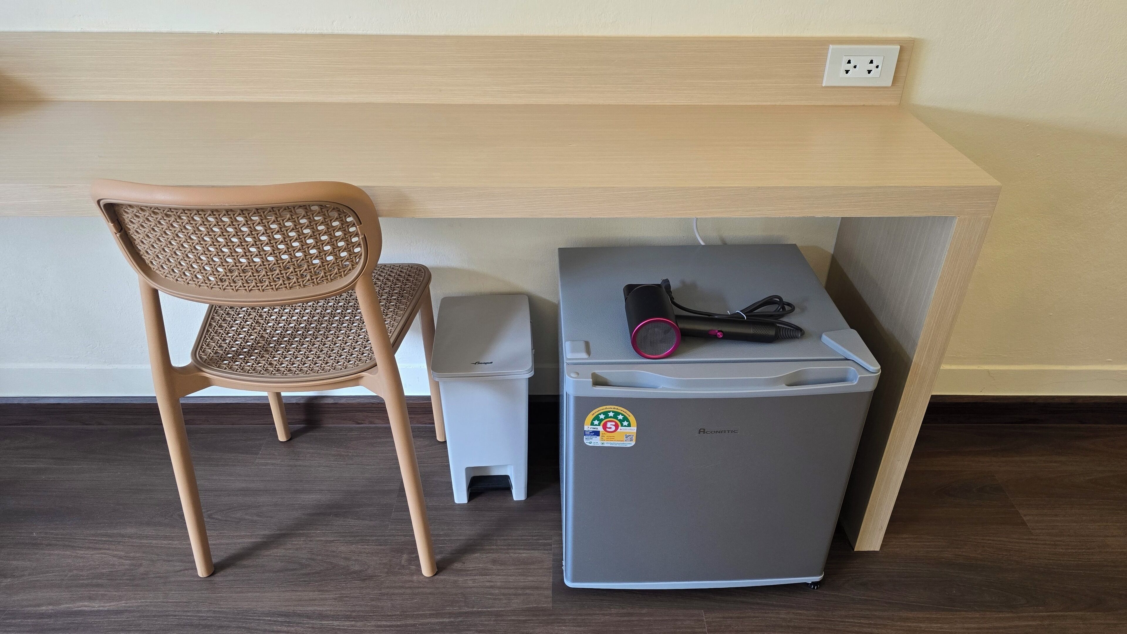 A compact kitchenette with a grey refrigerator, a small trash bin, and a chair under a wooden countertop.