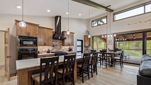 A modern kitchen with wooden cabinets, a large island with bar stools, and a dining area with a view. - New in Powder House! Pet Friendly, 4 Ensuites, Iconic Wraparound Deck (Lead)