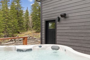 A hot tub outside a building with a wooden exterior, a black door, and a view of trees.