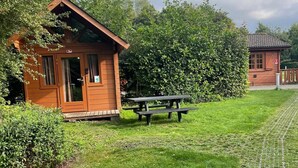 Una cabaña de madera con puerta y ventanas, una mesa de picnic y un área verde con una cerca de alambre.