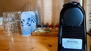 Una máquina de café Nespresso Magimatic sobre una mesa de madera con vasos de vidrio transparente, una taza con motivos azules y blancos, y una bandeja con cápsulas de café.