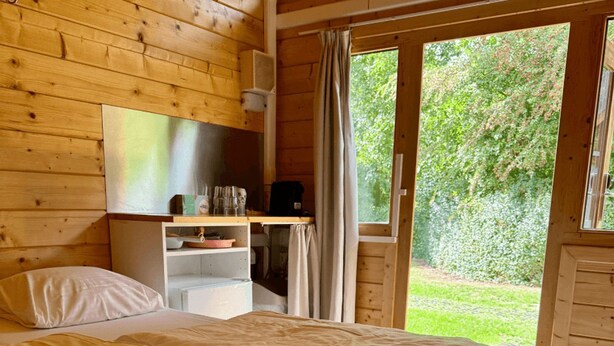 Un dormitorio de cabaña de madera con una cama, una mesita con objetos y vistas al verdor exterior.