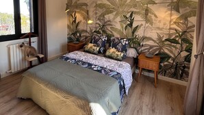 A bedroom with a bed, a ceiling fan, a radiator, a window with curtains, and a wall with a tropical plant mural.
